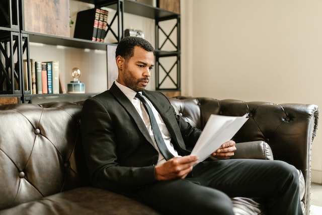 a landlord in a black suit sits on a brown leather couch and reviews the terms of a lease agreement prior to drafting an eviction notice for their renter